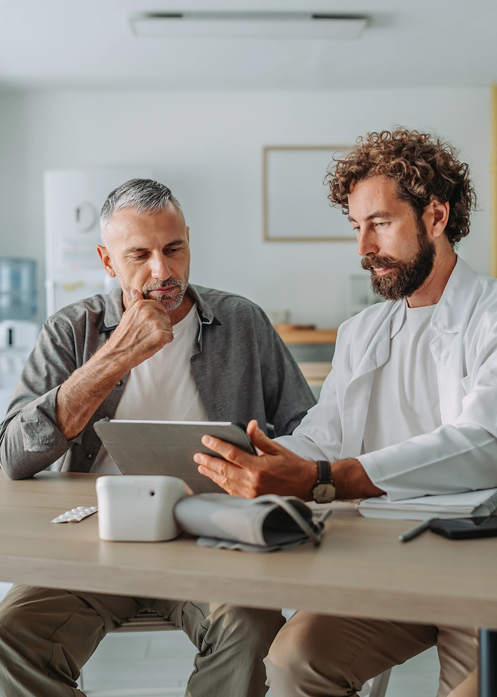 Médecin homme tenant un iPad dans ses mains en direction de son patient. Un tensiomètre est posé sur le bureau du cabinet médical.